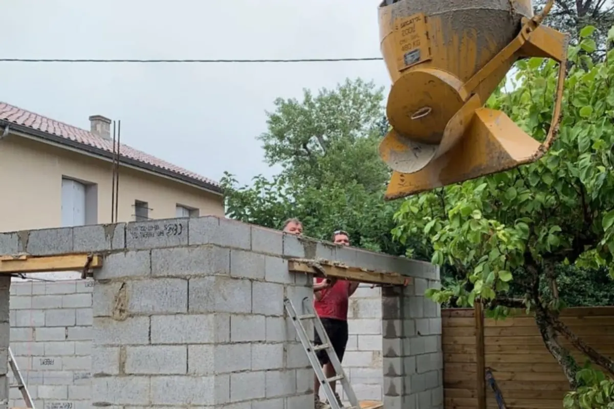 Maçons montant un mur en parpaing, benne à béton suspendue à la grue au-dessus du chantier