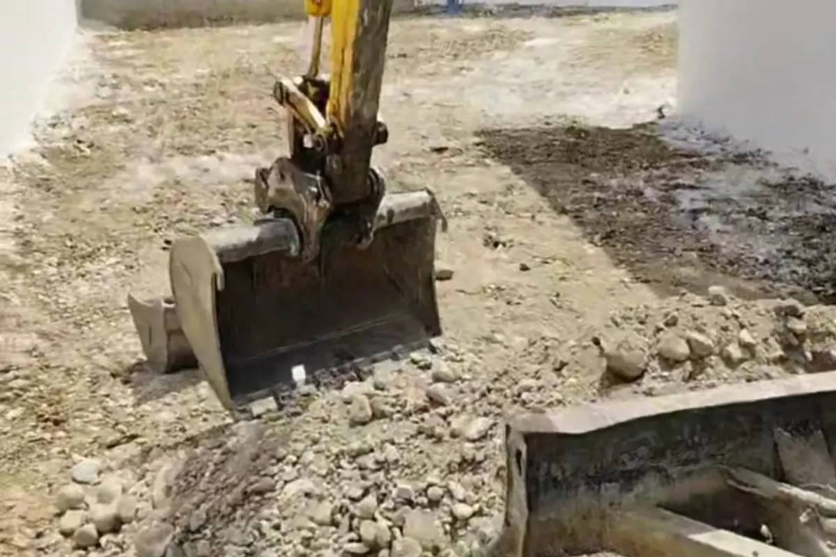 Godet de mini-pelle creusant une tranchée dans un terrain caillouteux, à l’intérieur d’un mur de clôture
